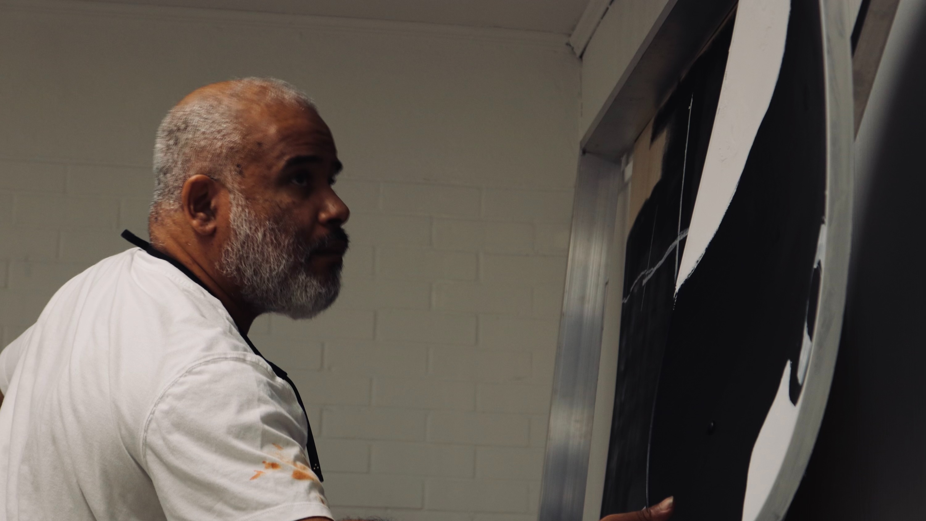 Marcus Jansen in a paint-stained white t-shirt working closely on a large black and white canvas in his studio, photographed in profile.
