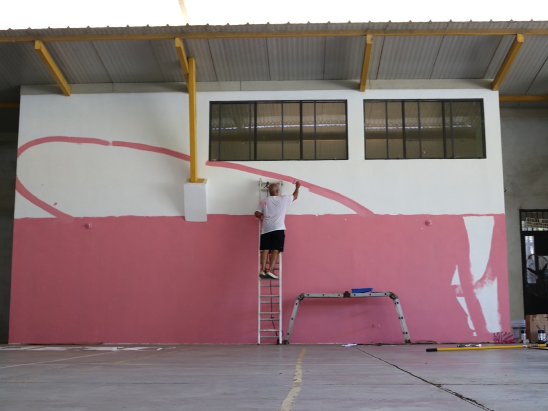 A man on a tall ladder paints a large pink geometric shape onto a high, white indoor warehouse wall under a yellow metal roof structure.
