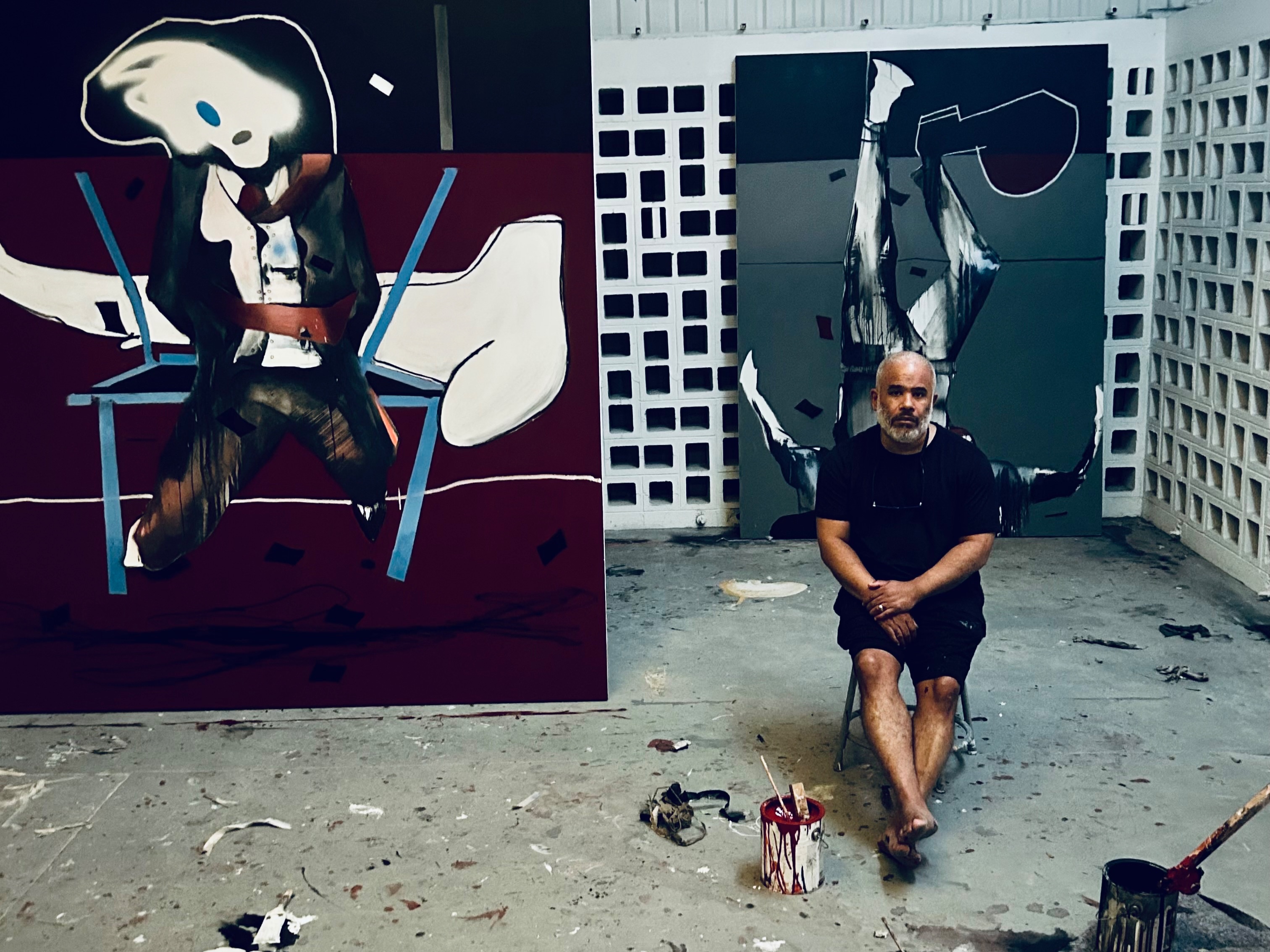 Marcus Jansen sits barefoot on a stool in a paint-spattered concrete studio, flanked by two large, dark surrealist paintings depicting stylized figures.